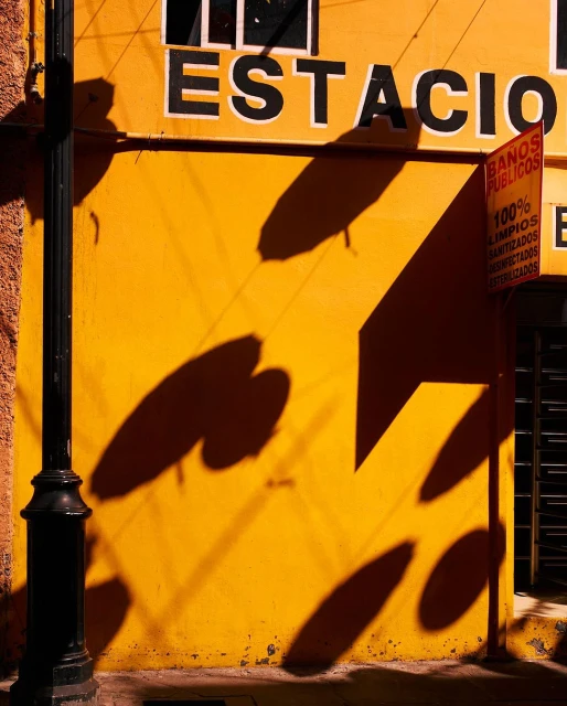 Bright yellow building facade with black lettering, shadows, and a black lamppost against a textured brick wall on a sunny day.