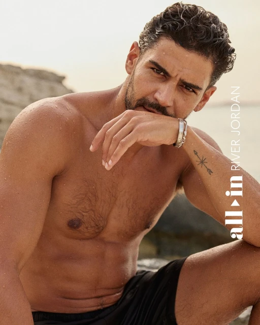 Portrait of a muscular adult male with a beard, wearing black shorts and silver bracelets, sitting outdoors in front of a neutral sandy background.