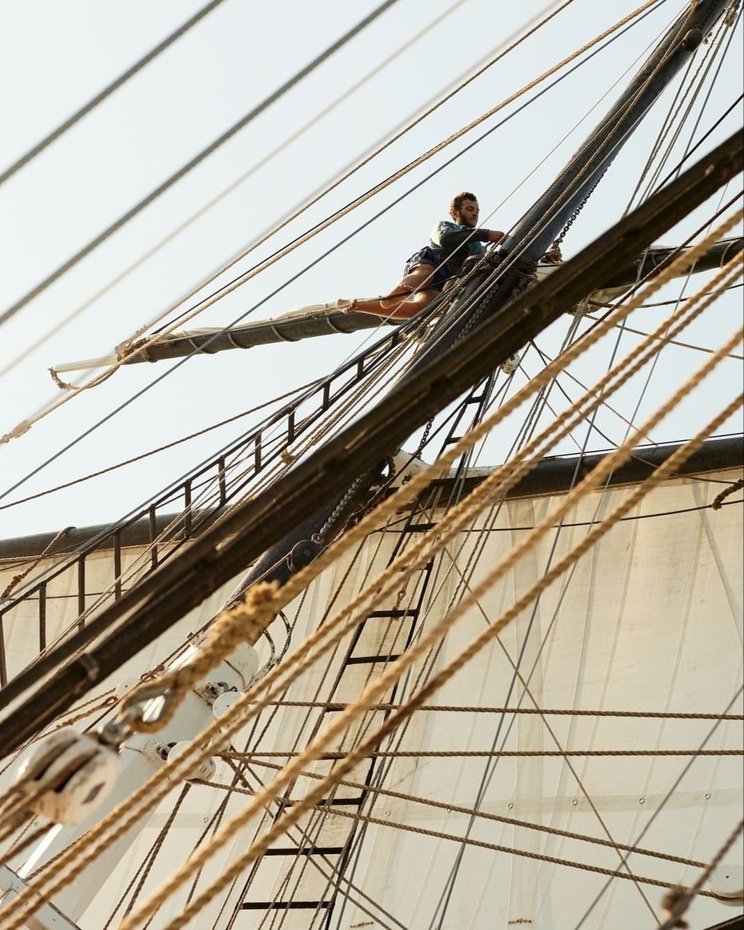 A man balances on the rigging of a tall ship, showcasing acrobatic skill and athleticism against a backdrop of ropes, sails, and sky.