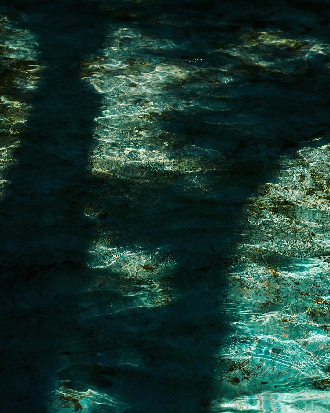Abstract close-up of turquoise water with light shining through, creating a rippled effect, with dark shadows in the background.