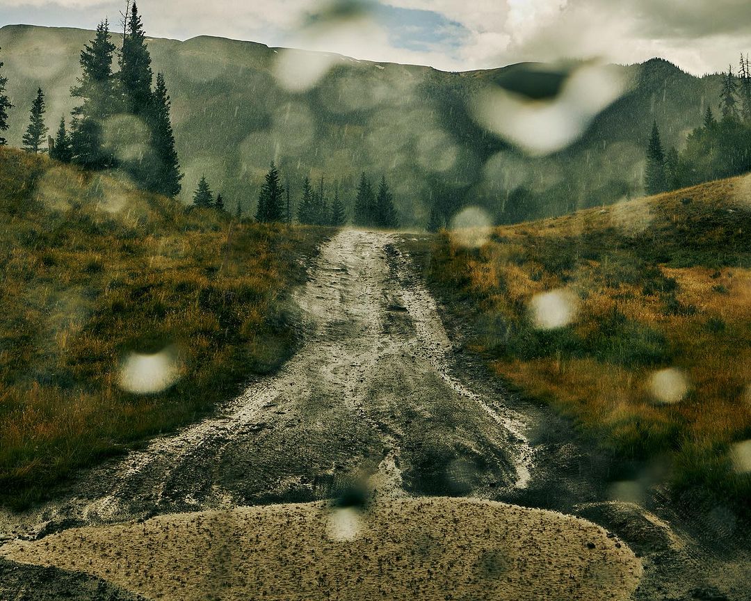 Scenic mountain landscape view through rain-speckled window. A dirt road stretches into the distance, surrounded by tall evergreen trees.