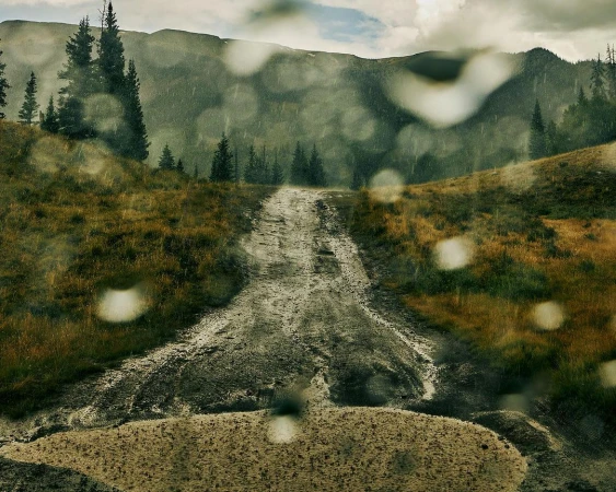 Scenic mountain landscape view through rain-speckled window. A dirt road stretches into the distance, surrounded by tall evergreen trees.