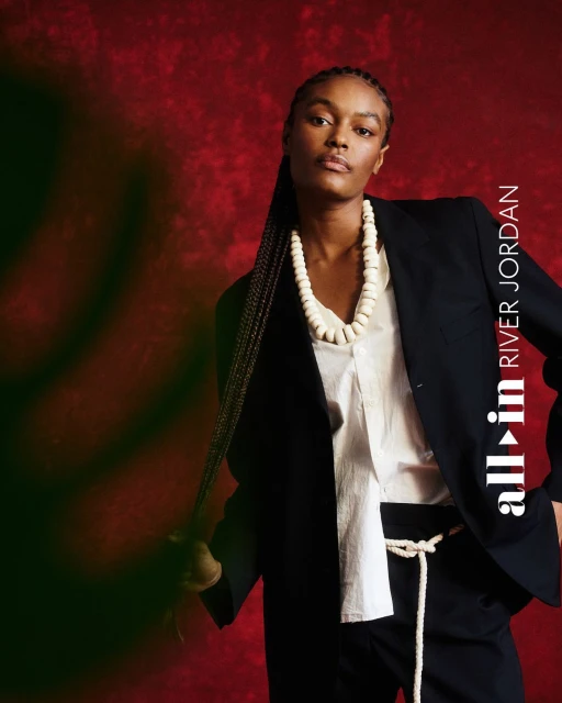 Portrait of Black woman in blazer against a red backdrop, wearing large bead necklace and rope belt, holding her braided hair.
