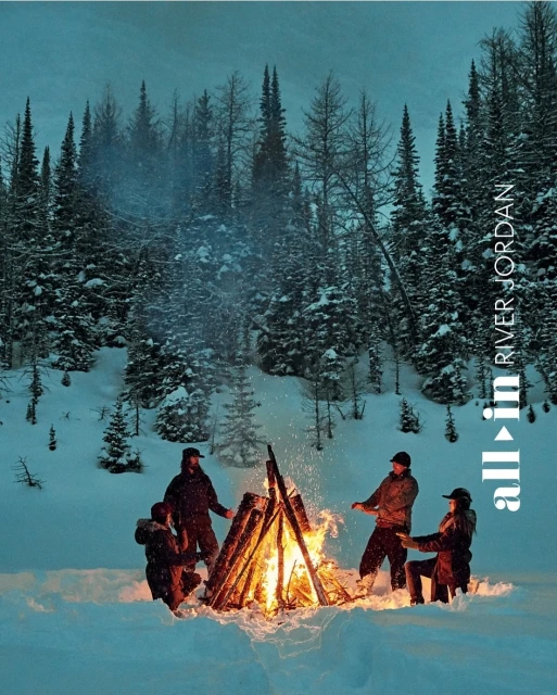 Group of five people gathering around a campfire in a snow-covered forest, with tall pine trees and a cold, serene winter scene.