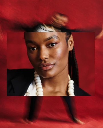 Portrait of a woman with braided hair wearing a beaded necklace and dark blazer against a textured red background