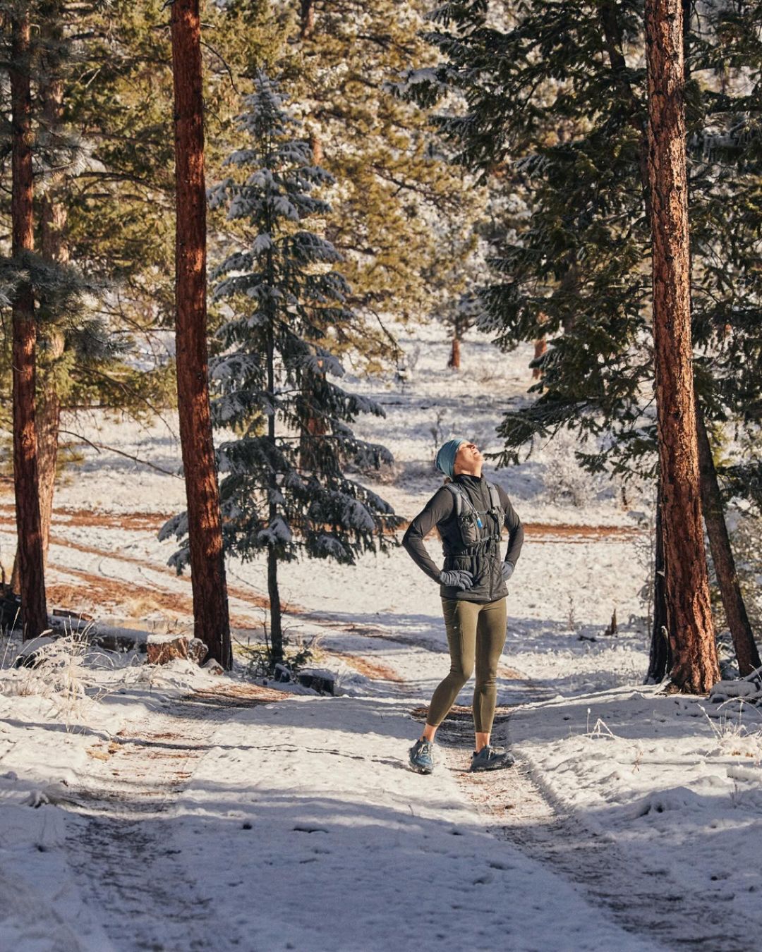 A woman stands on a snowy path in a forest, wearing athletic clothing and a head covering, looking up with hands on her hips on a sunny day.