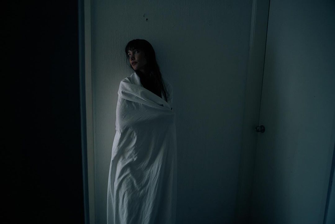 Woman wrapped in a white sheet standing in a room. Dark lighting and neutral colors create a somber mood.