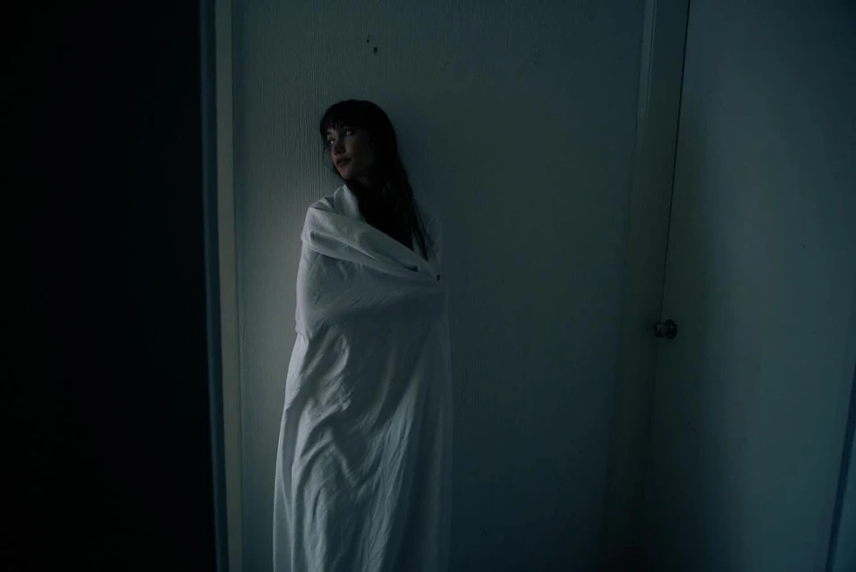 Woman wrapped in a white sheet standing in a room. Dark lighting and neutral colors create a somber mood.