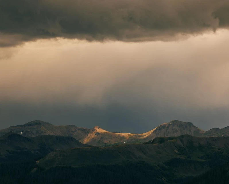 Dramatic mountain landscape with sunlit peaks under dark clouds, creating a serene yet powerful vista of nature's grandeur.