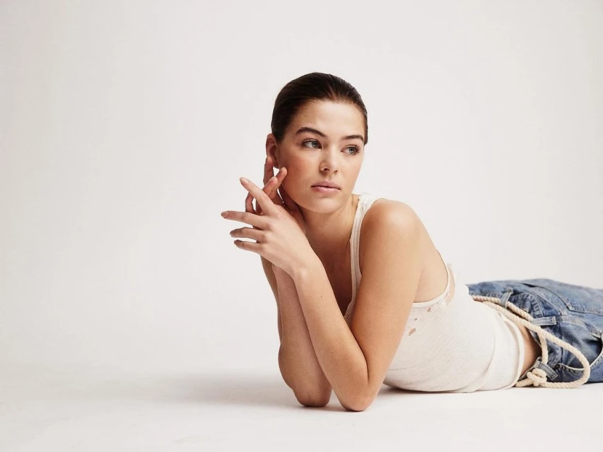 Portrait of a woman in casual clothes lying on her side in a studio setting with white seamless background, lit by soft natural light.