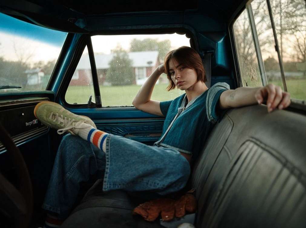 Woman in denim outfit sits inside vintage blue truck in a rural setting, wearing sneakers and striped socks, looking at the camera.