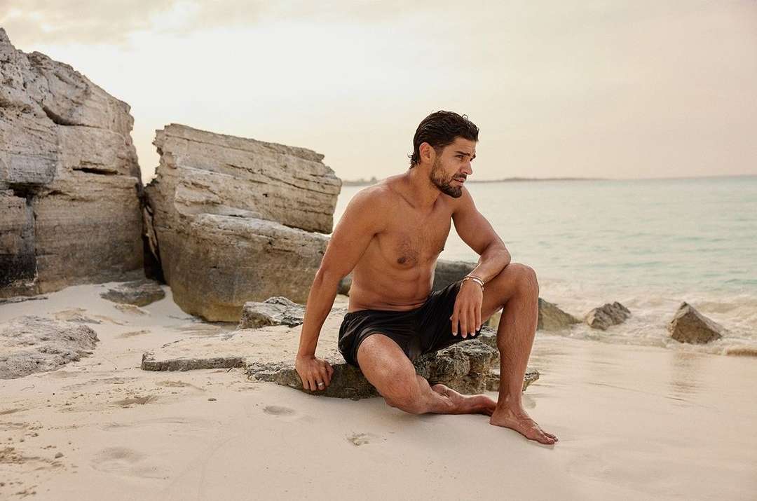 Adult male with bare chest and black shorts sits on a rock formation on a sandy beach with ocean and sky visible in the background.