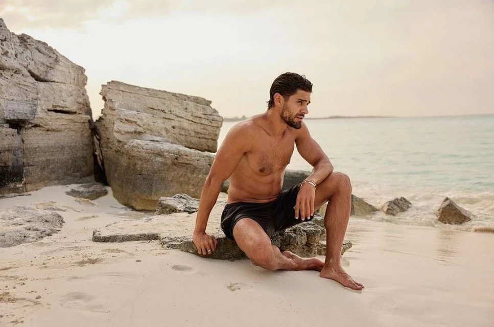 Adult male with bare chest and black shorts sits on a rock formation on a sandy beach with ocean and sky visible in the background.