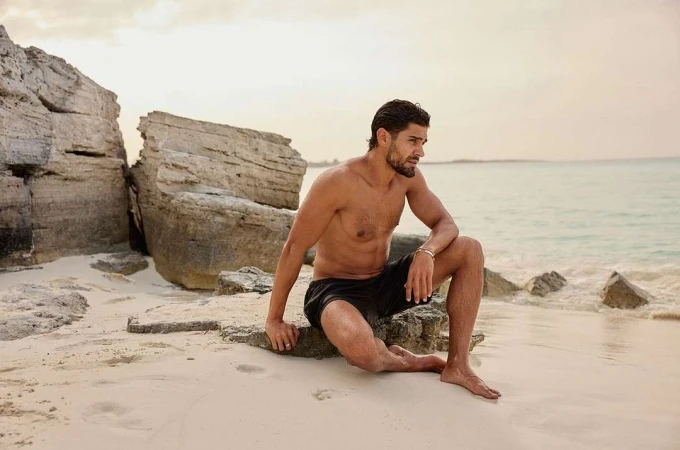 Shirtless man sitting on a rocky outcropping on a sandy beach looking out toward the ocean.