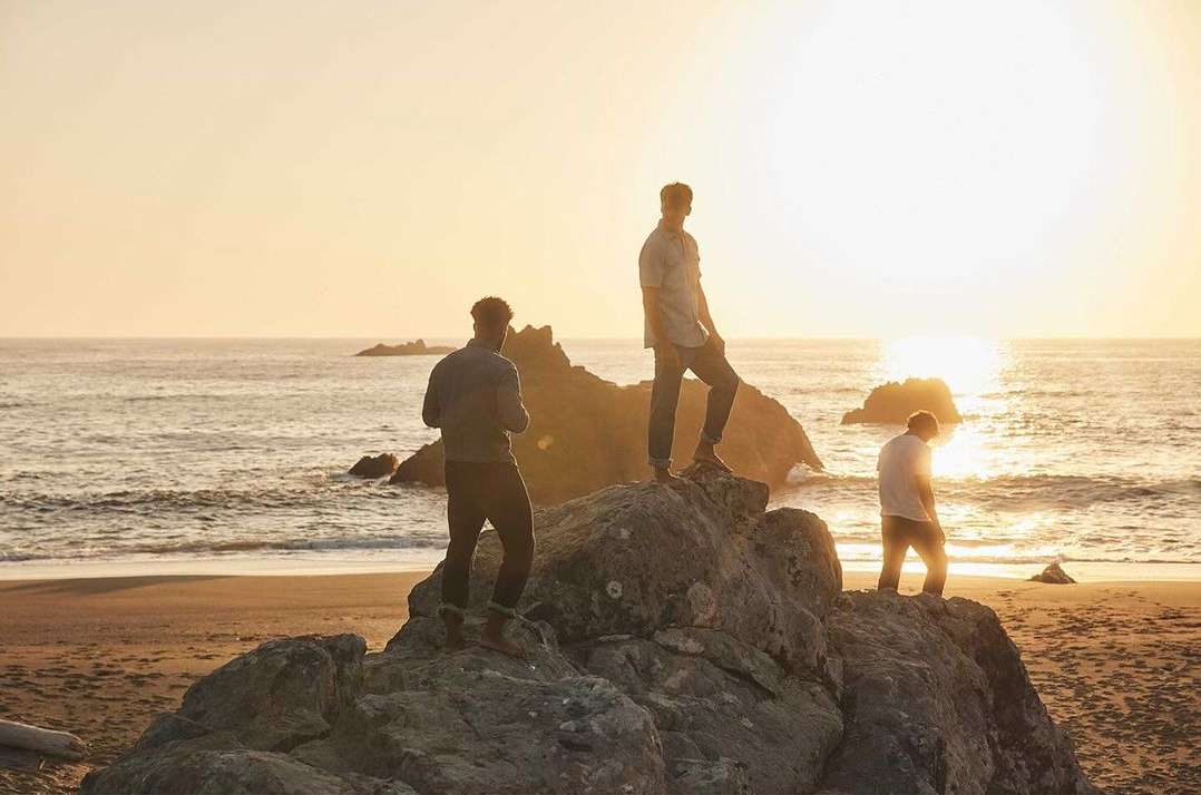 Three adult males stand on rock formations overlooking the ocean at sunset, enjoying the beach landscape and golden light.