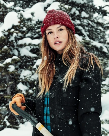 Attractive woman in winter clothing outdoors wearing a knit hat with a snow-covered evergreen background, looking at the camera.
