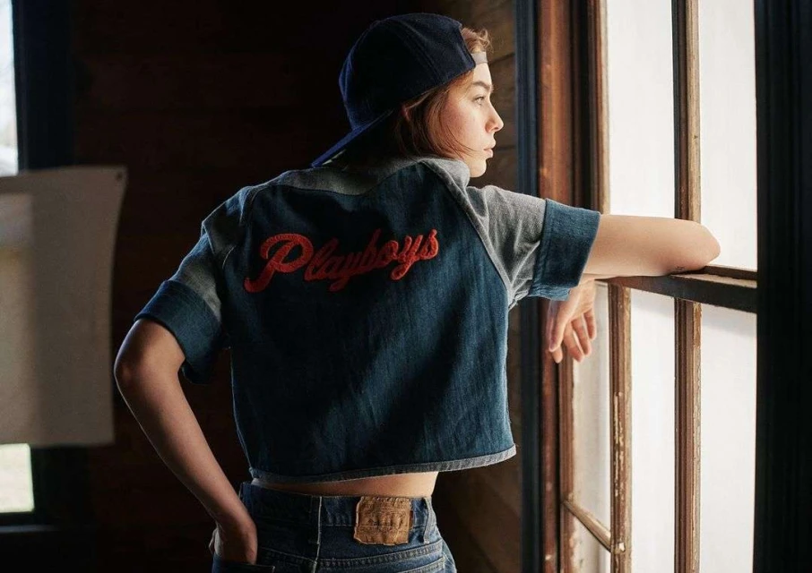 Young woman wearing a cap and denim jacket with "Playboys" on the back, looking out a window in an indoor setting.