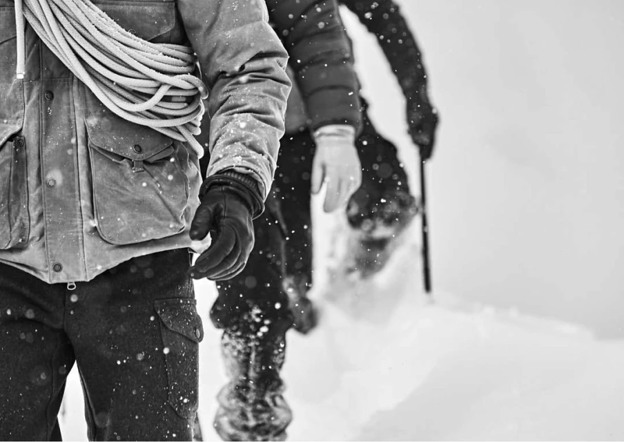 Close-up of two people in winter jackets and gloves walking through a snow-covered landscape. One person carries a coiled rope over their shoulder.