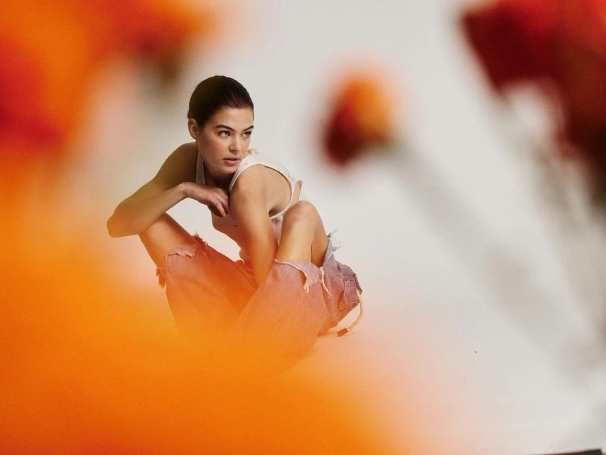 Young adult woman in torn jeans sits in a photo studio setting, partially obscured by a blurry orange foreground.