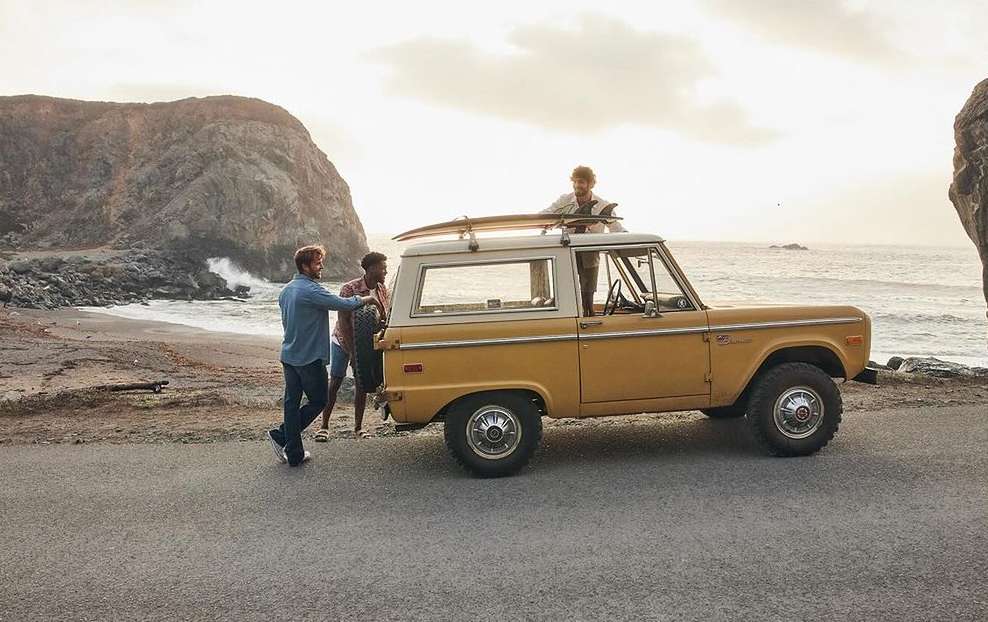 A vintage yellow and white car parked on a coastal road with three men, one on the car roof with a surfboard, ocean backdrop.