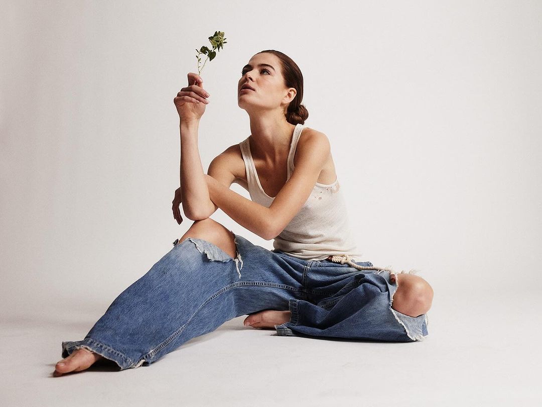 Woman in ripped jeans sits on the floor and holds a sprig of green leaves. She is in a photo studio in front of a white background.