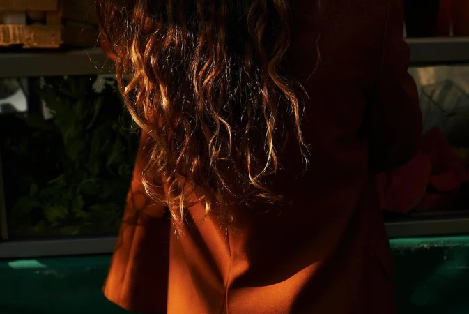 Rear view of a person with long curly blonde and brown hair wearing a rust-colored jacket.