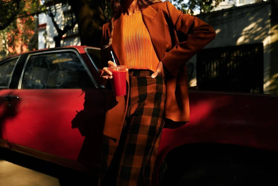 A woman in a blazer holds a red drink next to a red car in the city, bathed in warm sunlight. She is wearing plaid pants.