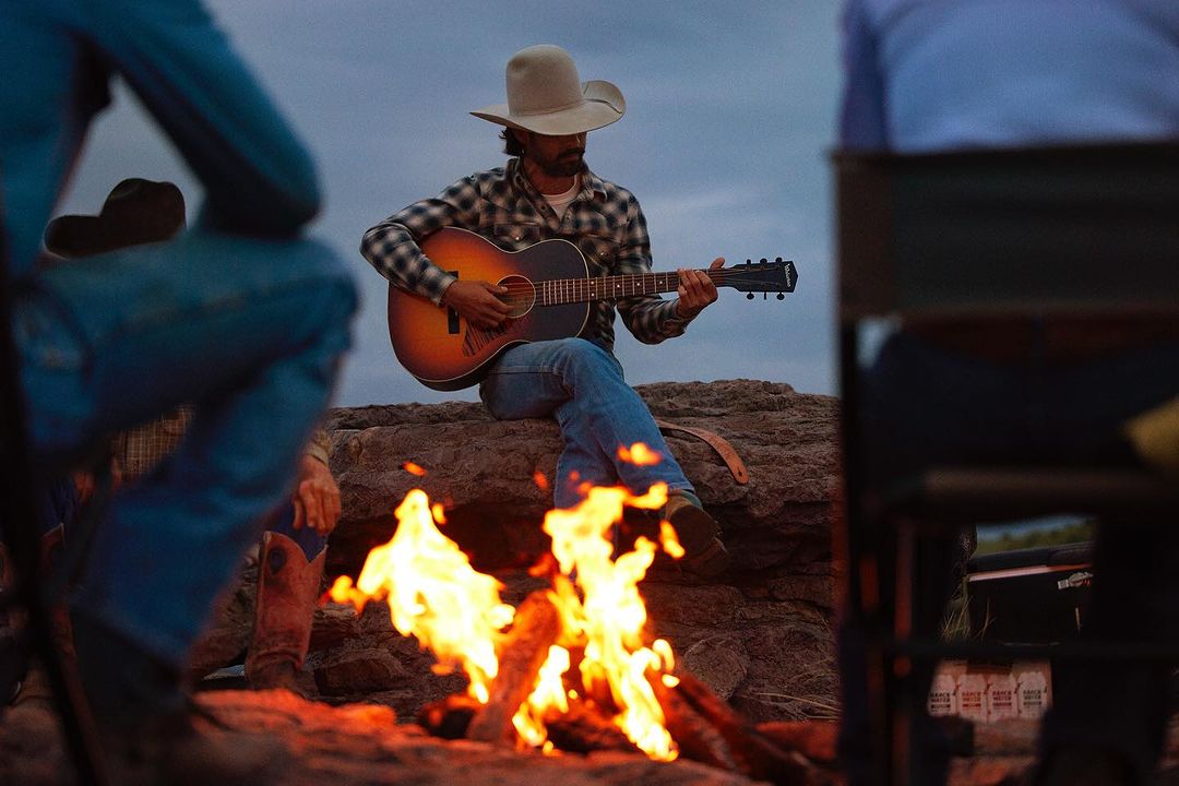 Cowboy plays guitar by a campfire on a rocky outcrop at dusk, wearing a cowboy hat, jeans, and plaid shirt. Group of people around.
