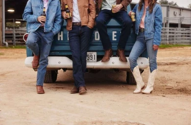 Four people leaning against the tailgate of a blue pickup truck holding beer bottles, wearing denim and boots.