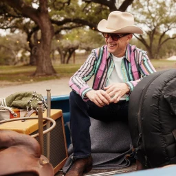 Smiling man in a cowboy hat and striped shirt sitting in a pickup truck bed outdoors by River Jordan.