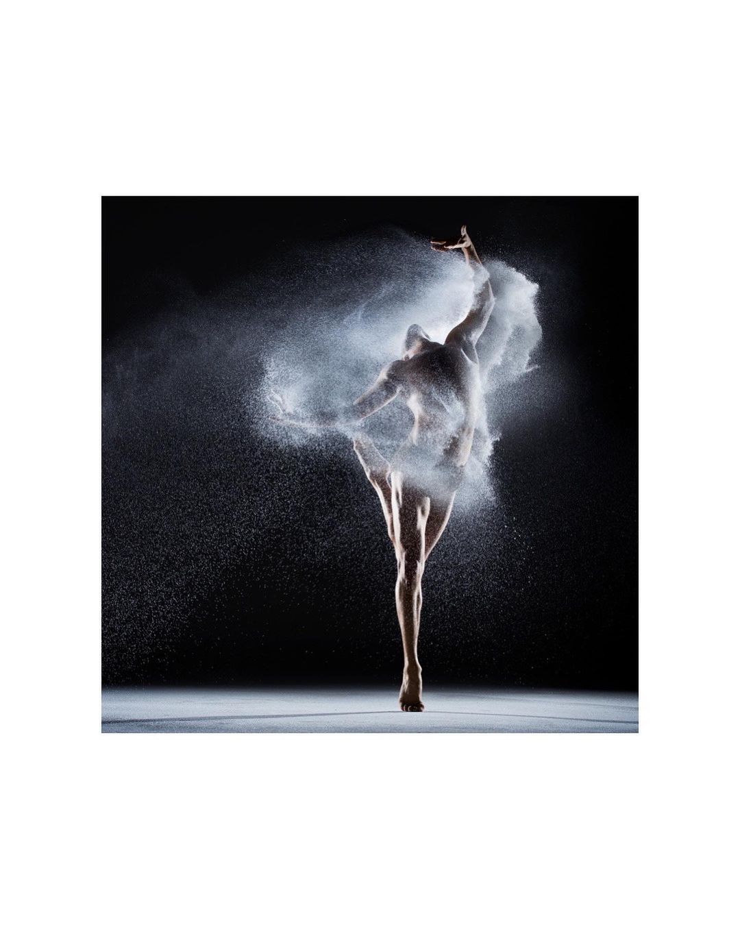 Artistic studio shot of a ballet dancer in a dynamic pose surrounded by a cloud of white powder against a dark background.
