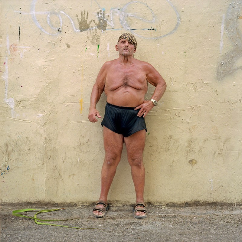 Full shot of a muscular, shirtless, adult male wearing a headscarf, shorts, and sandals posing in front of a beige graffiti wall outdoors.