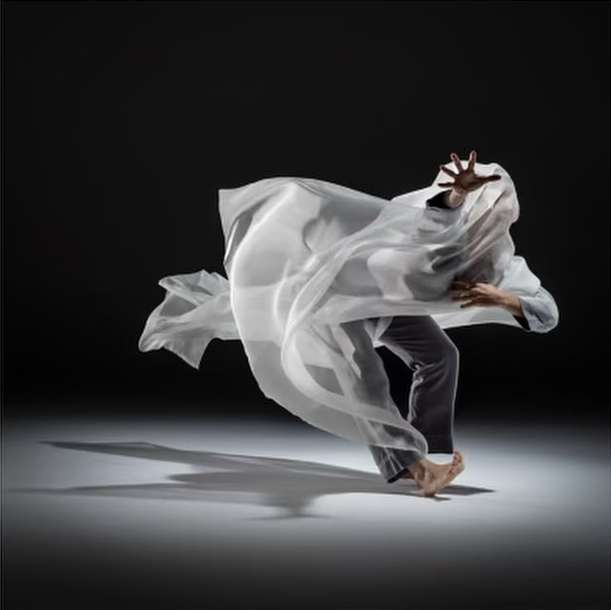 Person dancing with flowing white fabric in a dynamic studio shot against a dark backdrop, emphasizing movement and light.