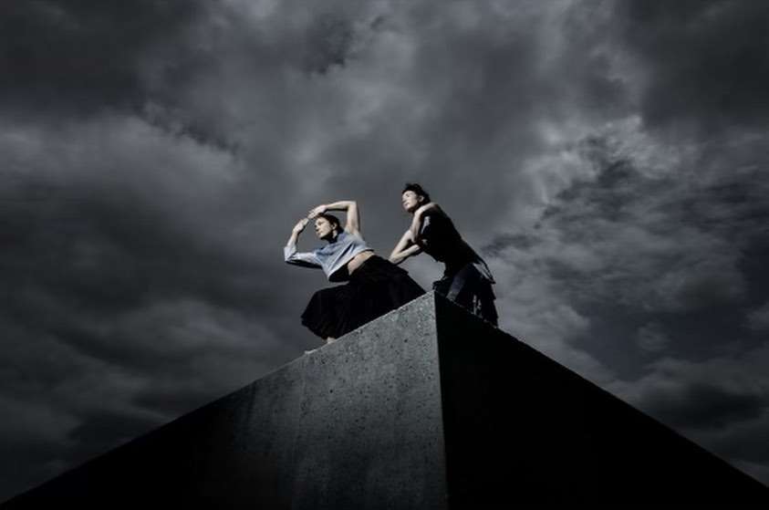 Two women strike a pose on a concrete block under a dramatic cloudy sky, embodying movement and strength against a stark background.