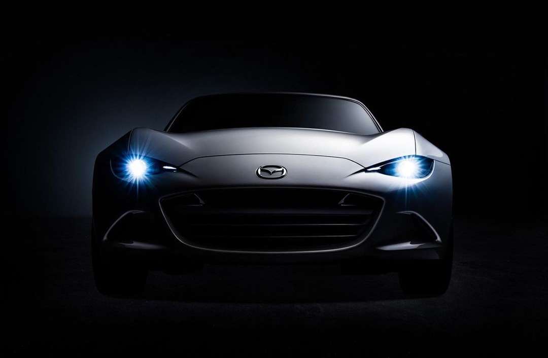 Sleek silver Mazda car with its headlights on, positioned in front of a dark background, showcasing its modern automotive design.