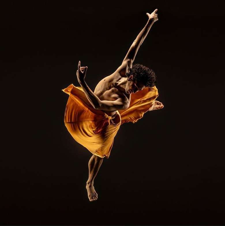 A dark-haired male dancer with a partially bare torso leaps in a studio, wearing an orange flowing garment against a black background.