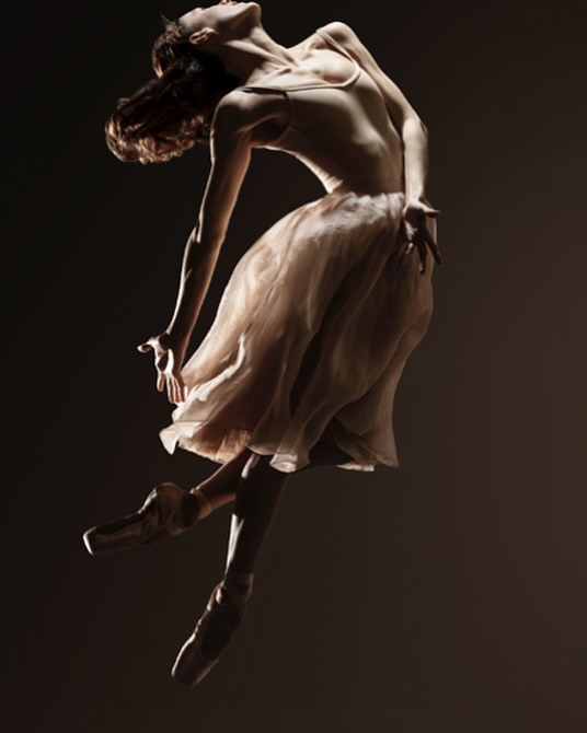 A ballerina performs an elegant dance in a dramatic pose, leaping with her back arched, bathed in warm, focused studio lighting.