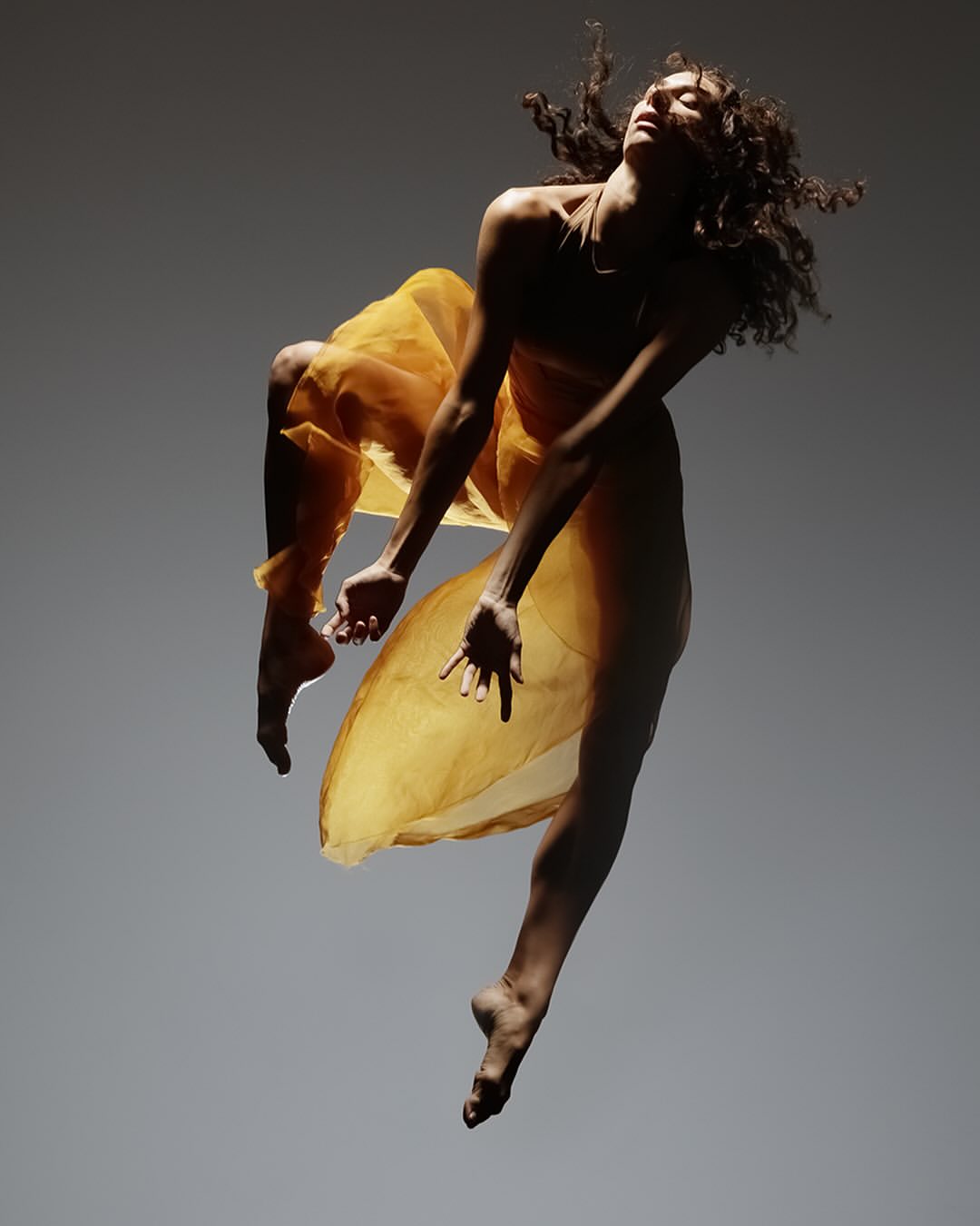 Woman in mid-air, gracefully suspended in a yellow dress, showcasing dance and movement against a grey backdrop in a studio setting.