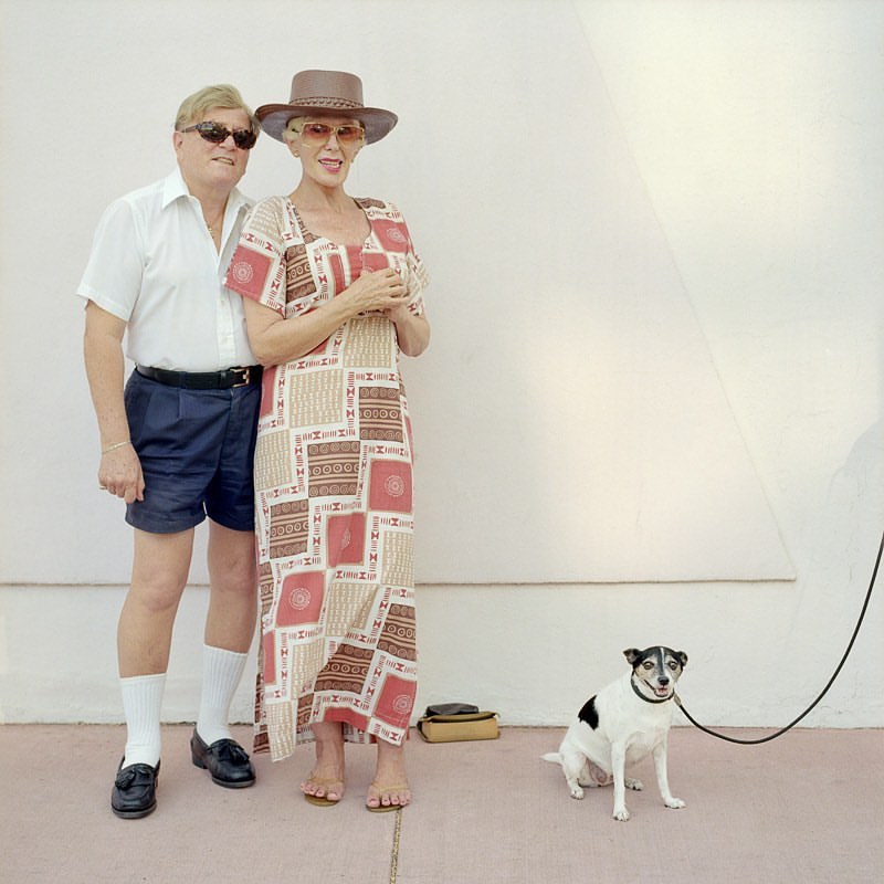 Elderly couple poses with a small dog on a leash in front of a white wall. The woman wears a long patterned dress and the man wears shorts.
