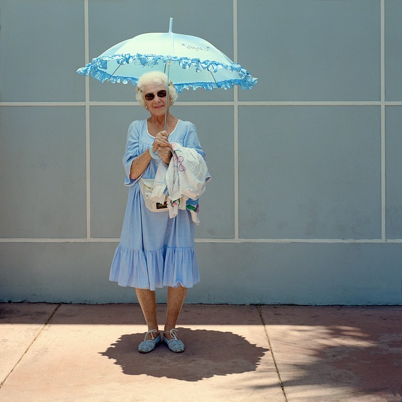 Elderly woman in a blue dress stands in the shade of a matching blue umbrella holding a white cloth bag on a sunny day.