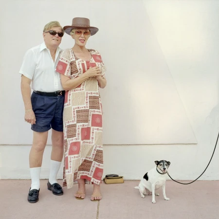 An older couple posing outdoors with a small black and white dog on a leash against a plain wall.