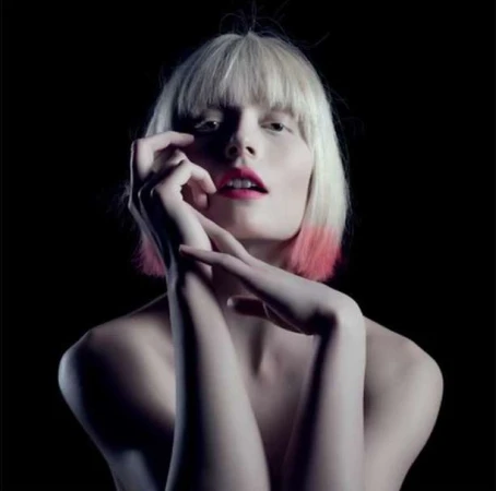 Portrait of a blonde woman with pink-tipped hair and bright pink lipstick posing with her hands near her face against a black background.