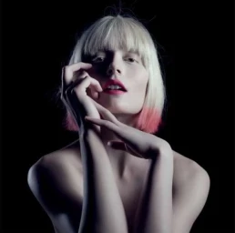 Portrait of a blonde woman with pink-tipped hair and bright pink lipstick posing with her hands near her face against a black background.