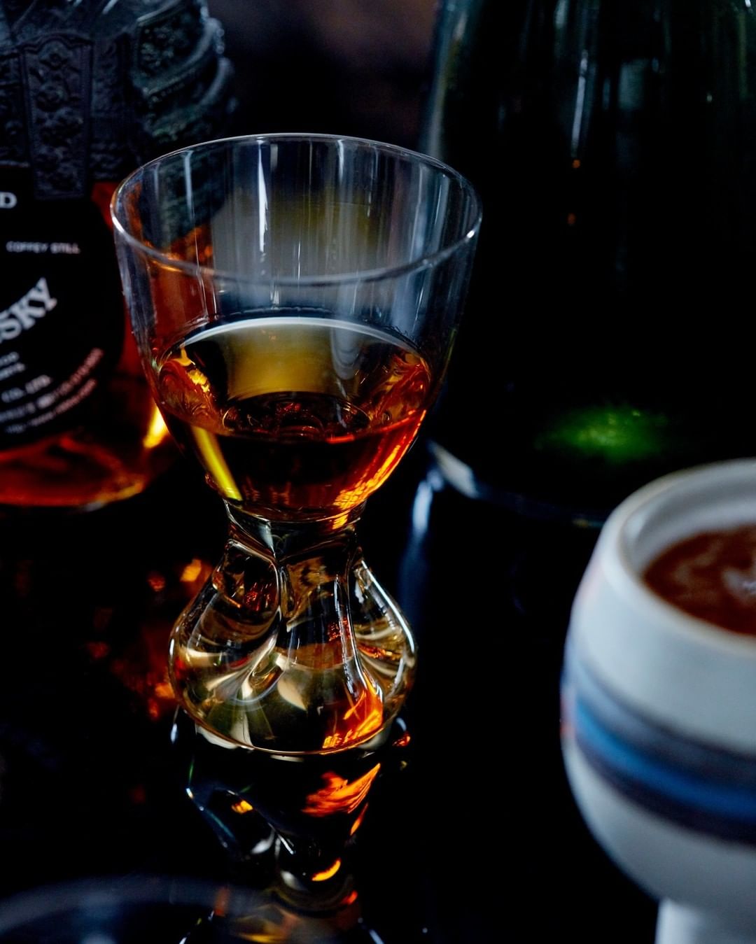 A glass with a dark liquid sits on a reflective surface, accompanied by liquor bottles and a small patterned cup in the background.
