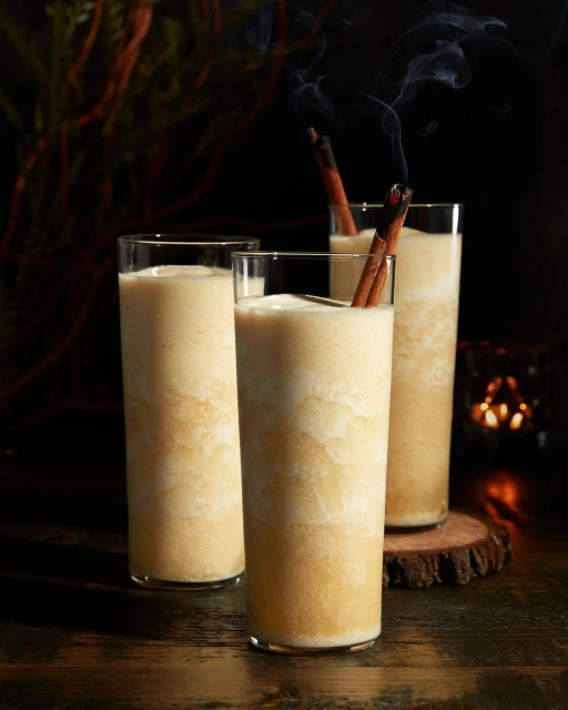 Three glasses of a creamy beverage with cinnamon sticks, displayed against a dark backdrop with foliage and warm candle-like lighting.