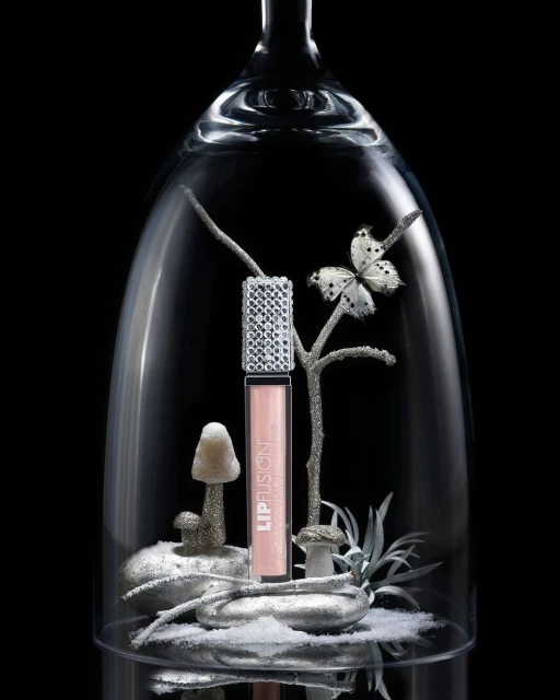 Lip gloss product shot featuring a tube of lip gloss under a glass dome with mushrooms and a butterfly on a black background.