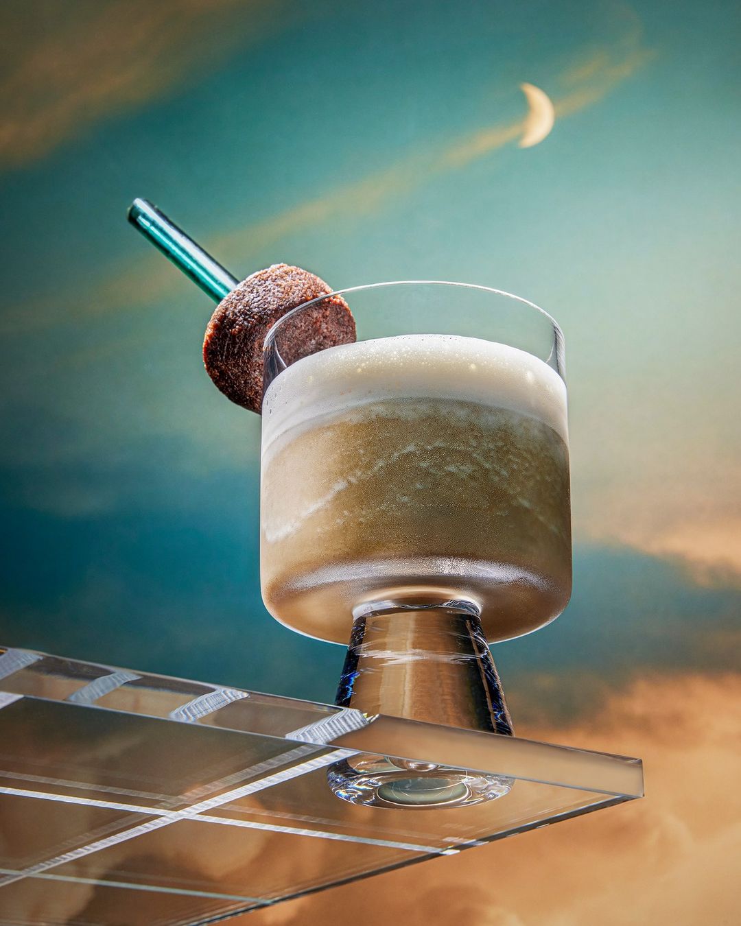 A cocktail in a stemmed glass with a straw and spherical garnish sits on a reflective surface with a colorful sky background.