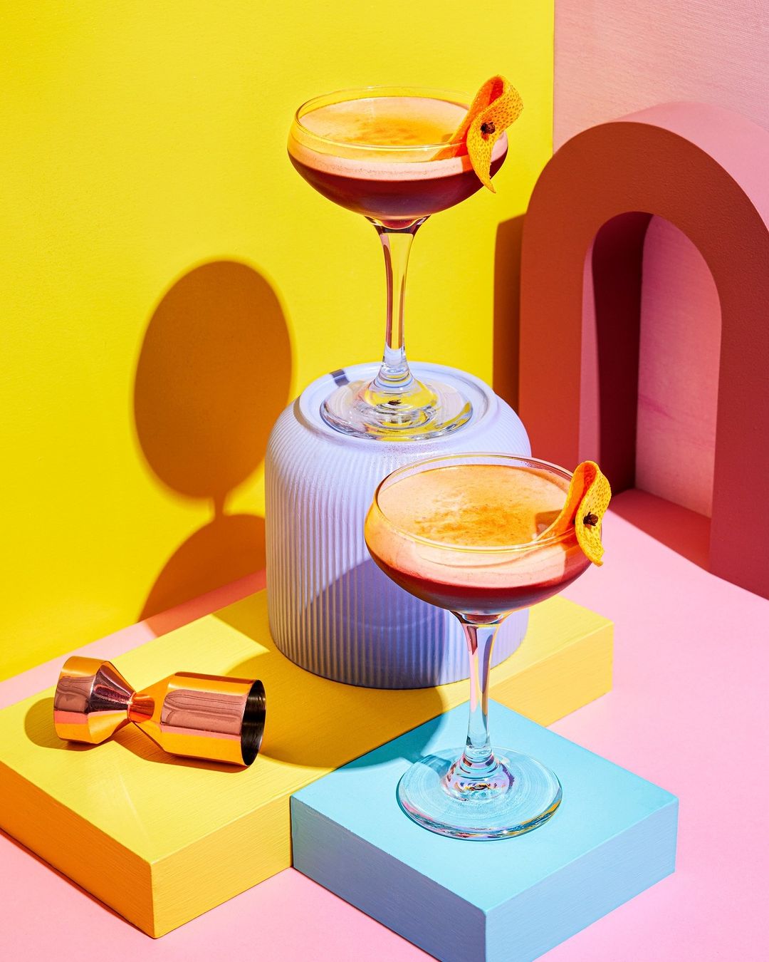 Two cocktails on colorful blocks with a copper jigger, capturing a vibrant and modern aesthetic in a studio setting with dynamic lighting.