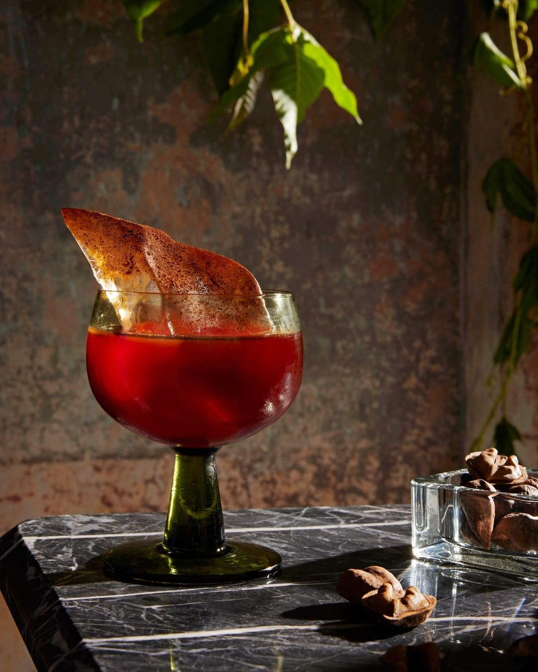 A cocktail in a green stemmed glass sits on a marble table with chocolate pieces, against a textured wall with green vines.