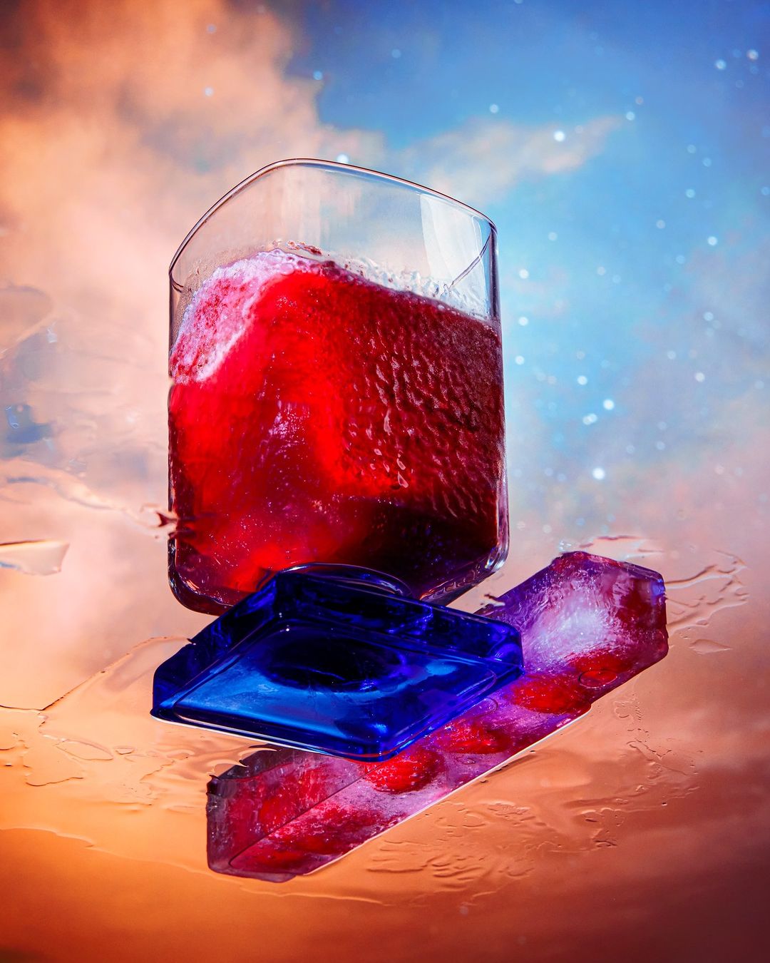 Artful still life of a glass filled with red liquid atop reflective surfaces, with a backdrop of a dreamy sky.